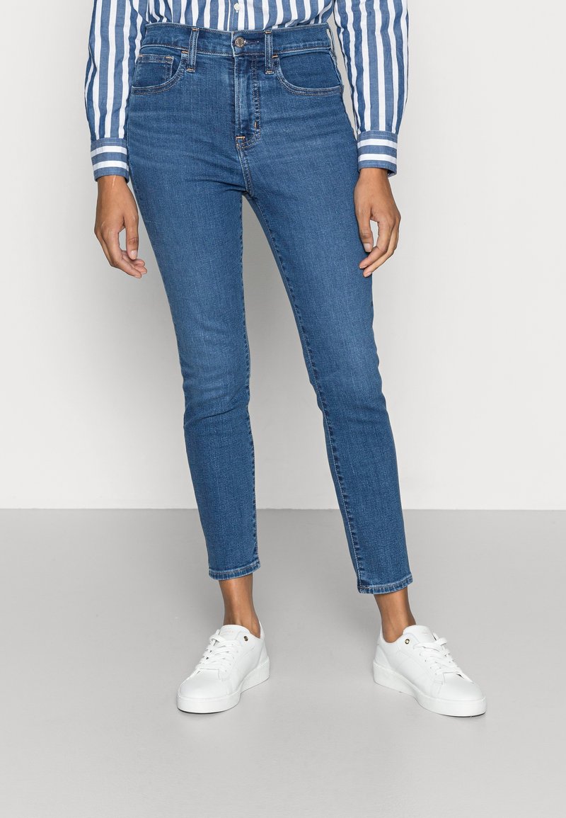 Jean en denim bleu foncé coupe slim, taille haute, cinq poches, avec des coutures discrètes, associé à des baskets blanches.