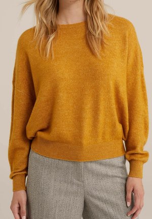 Vrouw draagt een mosterdgele gebreide trui met geribbelde manchetten en zoom, gecombineerd met grijze visgraatbroek, staand tegen een neutrale achtergrond.