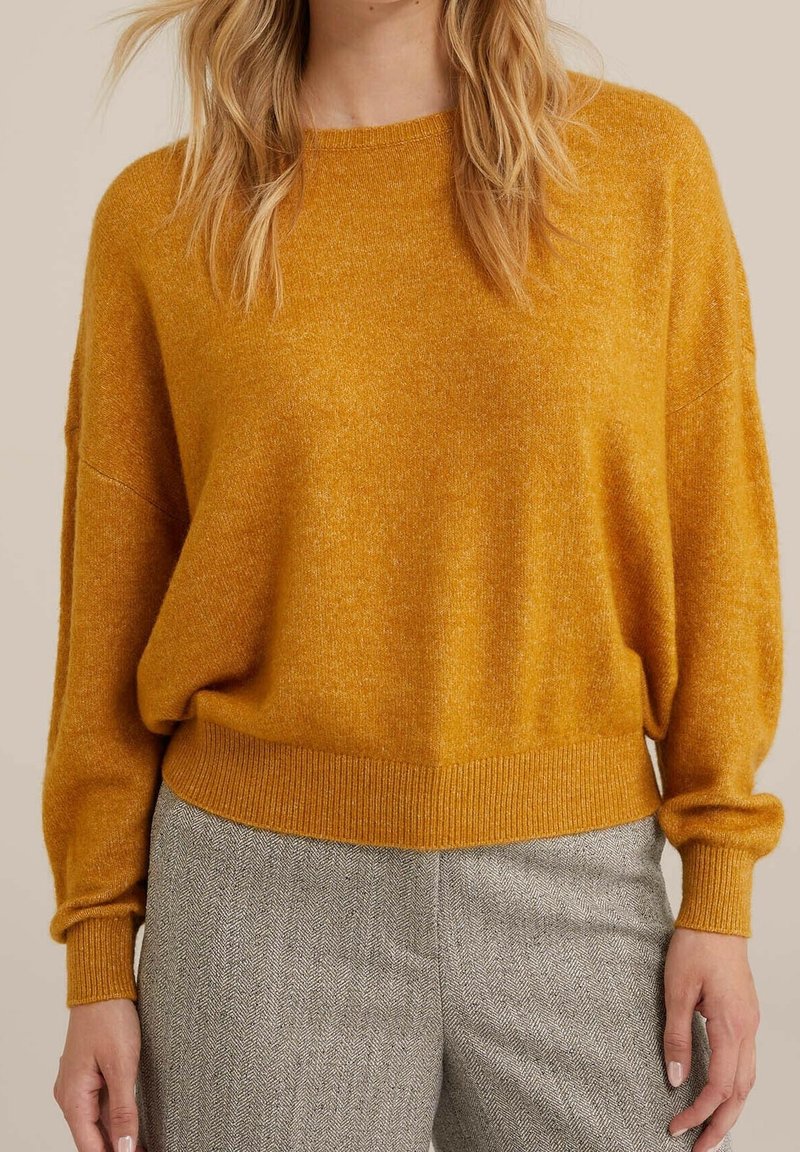 Femme portant un pull en maille jaune moutarde avec des poignets et un ourlet côtelés, associée à un pantalon à chevrons gris, debout devant un fond neutre.