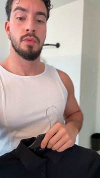 Polo nera su una gruccia nera tenuta da una persona con una canottiera bianca a coste; gancio di filo visibile. Sfondo neutro con illuminazione morbida.