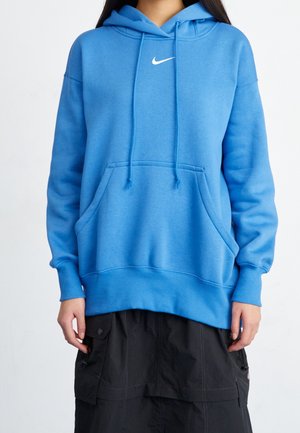 Henkilö jolla on kirkuvan sininen Nike-huppari, jossa on etutasku ja kiristysnauhat, yhdistettynä mustaan cargo-tyyliseen hameeseen.