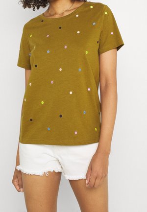 Vrouw draagt een olijfgroen T-shirt met korte mouwen met kleine kleurrijke stippen en witte versleten denimshorts, staand tegen een effen achtergrond.