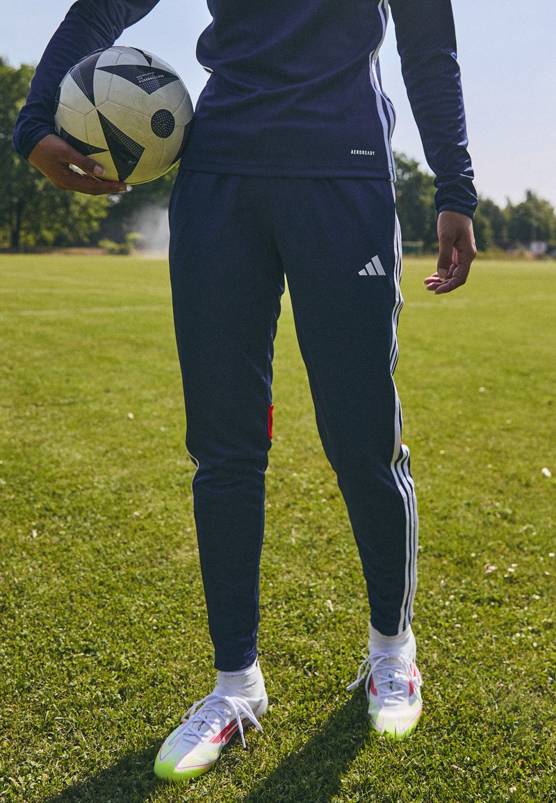 Adidas navy training pants with white stripes, paired with colorful soccer cleats. A black and white soccer ball is held in the left hand.