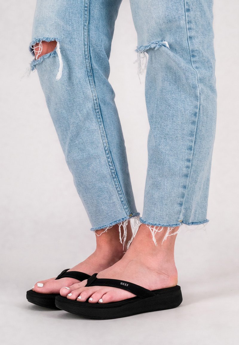 Black flip-flops with textured straps and cushioned soles, worn with light blue ripped jeans featuring raw hem and frayed edges.