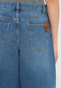 Los vaqueros de denim azul presentan una cintura alta, dos bolsillos traseros y un parche de cuero marrón cerca de la cintura. La tela tiene una textura de sarga clásica.