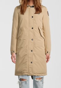 Beige parka med hætte, udstyret med sorte knapper, sidelommer og en let tekstureret overflade, båret over slidte lyseblå jeans.