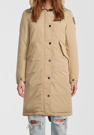 Parka beige avec une capuche, dotée de boutons noirs, de poches latérales et d'une surface légèrement texturée, portée sur un jean bleu clair déchiré.