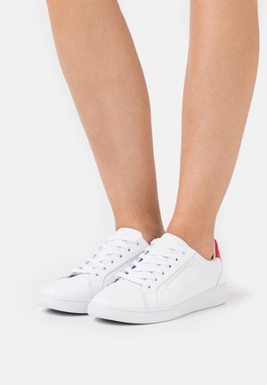 Baskets blanches à tige basse avec lacets et empiècements rouges au talon, portées sur des jambes nues devant un fond blanc.