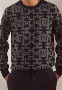 Maglione blu navy con un motivo geometrico grigio che presenta anelli e linee. Maniche e colletto a costine, realizzato in morbido tessuto a maglia.