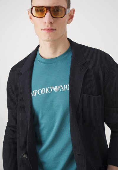 Juodas pilkšvas kardiganas su kišene, uždėtas ant melsvo marškinėlių su baltu logotipu. Modelis dėvi gintarinio atspalvio akinius.