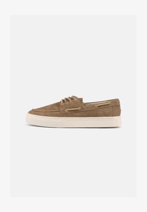 Zapato casual marrón de ante con cordones, suela de goma beige, ojales metálicos y costura moc toe, visto de lado sobre un fondo blanco.