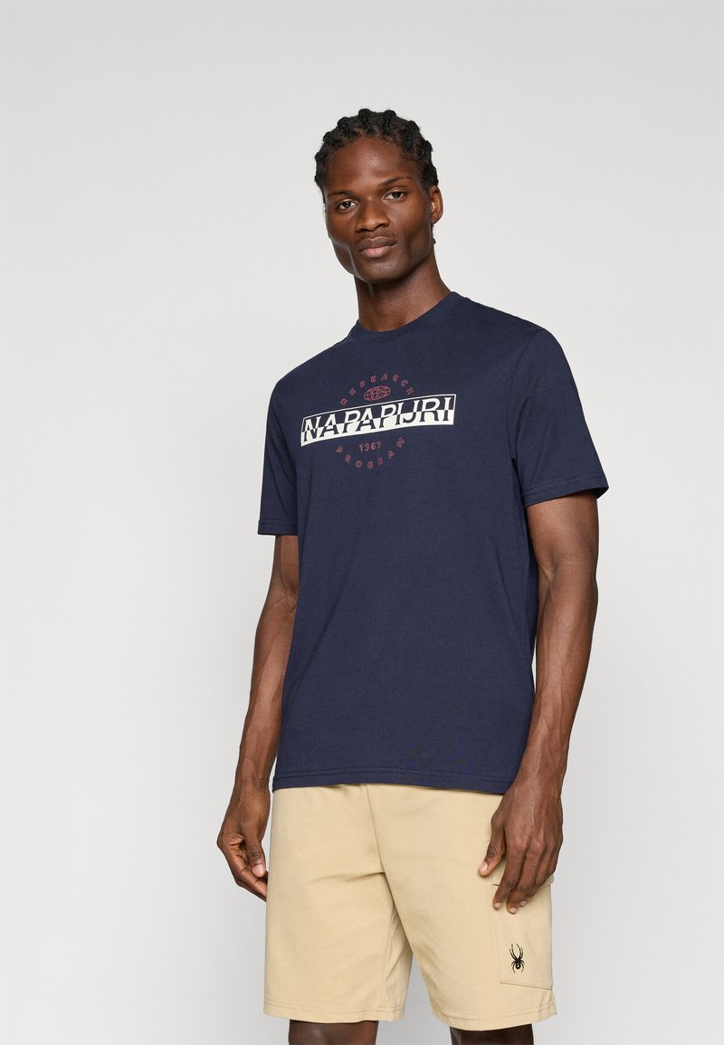 T-shirt en coton bleu marine avec le logo "NAPAPIJRI" en blanc et rouge, col rond, manches courtes, porté avec un short beige arborant un logo d'araignée sur la poche.