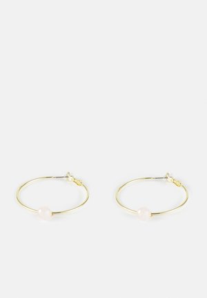 Boucles d'oreilles fines en forme d'anneau en or avec une petite perle ronde rose pâle sur chacune, maintenues par des fermoirs en plastique transparent sur un fond blanc.