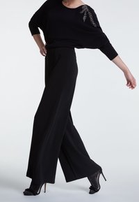Pull noir en maille avec un design drapé et des perles décoratives sur l'épaule, associé à un pantalon noir à jambe large. Escarpins noirs scintillants.