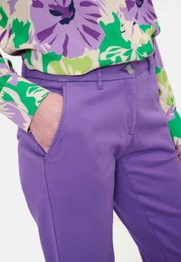 Pantalon en coton violet avec un devant plat, des poches inclinées et une fermeture par bouton. Associé à un chemisier vert à motif floral.