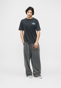 Νεαρός άνδρας στέκεται με τα χέρια στις τσέπες, φοράει ξεβαμμένο μαύρο μπλουζάκι με γραφικό σχέδιο, φαρδιά γκρι παντελόνια και λευκά αθλητικά παπούτσια, μπροστά σε απλό φόντο.