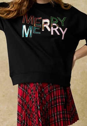 Personne portant un sweat-shirt noir avec le texte multicolore « MERRY MERRY » et une jupe plissée à carreaux rouges, bras plié à la taille.