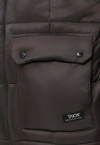 Sötétbarna puffkabát, amelyen patentos fedélzseb, vízszintes varrás és jelzett címke található a jobb alsó részen.