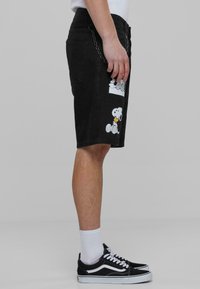 Crne traper kratke hlače s printovima Snoopyja i Charlieja Browna, s detaljem od lanca na pojasu, u kombinaciji s crnim tenisicama i bijelim čarapama.