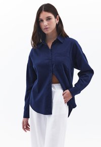 Marineblaue Button-Down-Bluse mit einer Brusttasche, lockerer Passform und langen Ärmeln, kombiniert mit weißen Hosen. Glatte Textur, minimalistisches Design.