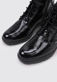 Botas de tobillo de piel de charol negra con punta redonda, que cuentan con un diseño de cordones y una superficie texturizada, montadas sobre una suela de goma baja.