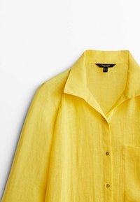 Camicia gialla in lino a maniche corte con colletto a punta, tasca frontale e chiusure a bottone. Tessuto strutturato con un aspetto leggero.