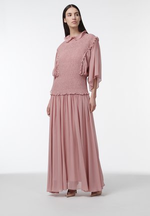 Mujer con un vestido largo rosa plisado con mangas con volantes y cuello alto, de pie frente a un fondo claro y liso.