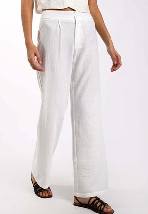 Pantalones de lino blanco de pierna ancha con pliegues frontales, cierre de botón y un acabado suave y texturizado. Llevados con sandalias negras de tiras.