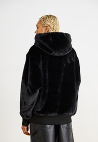 Černá umělá kožešinová mikina s kapucí, oversized design, texturovaný povrch, velká kapuce, žebrovan�ý lem a manžety. Zadní pohled bez viditelných zapínání nebo log.