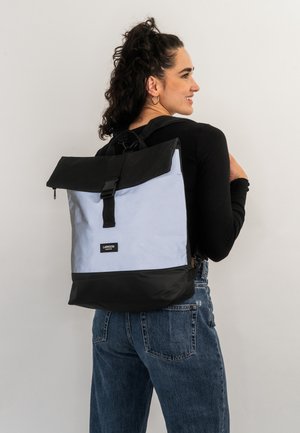 Sac à dos roulé bleu clair et noir avec des bretelles réglables, comportant une fermeture à boucle à l'avant et une étiquette logo en bas à l'avant.