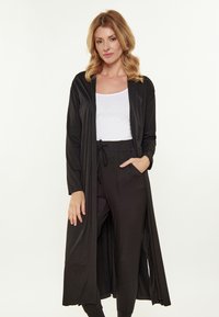 Schwarzer langer Cardigan aus weichem Stoff, mit lockerer Passform, offenem Vorderteil, langen Ärmeln und seitlichen Taschen. Kombiniert mit schwarzen Hosen und einem weißen Oberteil.