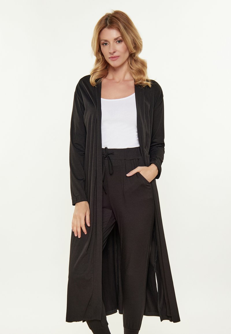 Schwarzer langer Cardigan aus weichem Stoff, mit lockerer Passform, offenem Vorderteil, langen Ärmeln und seitlichen Taschen. Kombiniert mit schwarzen Hosen und einem weißen Oberteil.