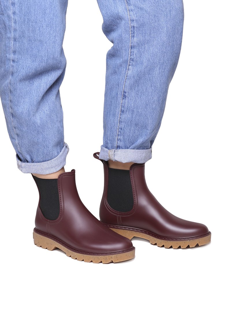 Botas Chelsea de goma burdeos con paneles elásticos negros, suelas texturizadas marrones y punta redondeada; combinadas con unos pantalones vaqueros azul claro remangados.