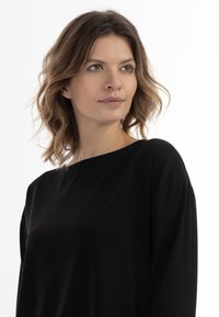 Musta pitkähihaine paita, jossa on veneen kaula-aukko, valuvakangas ja rento malli. Sujuva pinta ja minimalistinen muotoilu ilman näkyviä kuvioita.