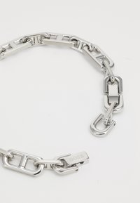 Silver metal chain bracelet featuring interlocking links, flat and rounded shapes, with a branded clasp detail. Minimalist design.