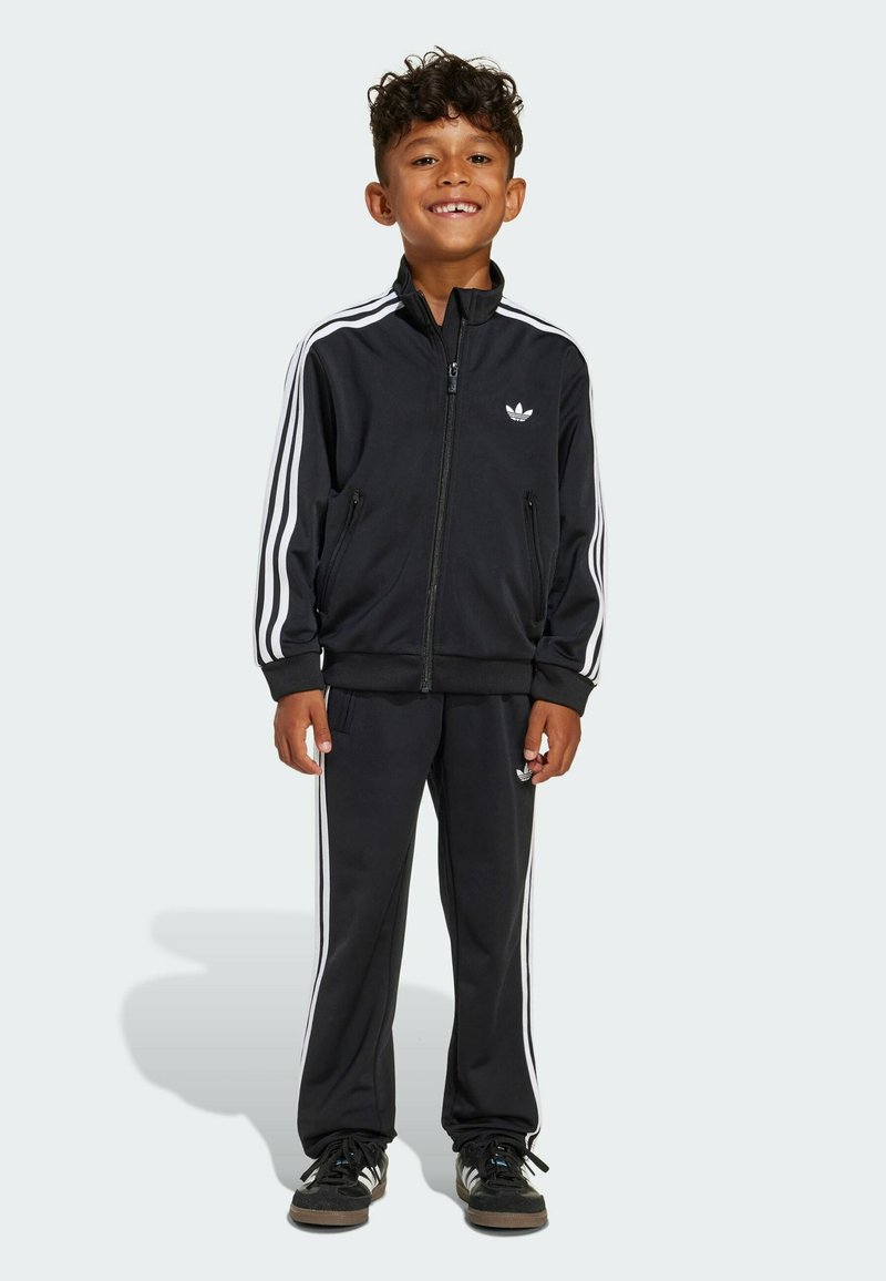 Smiling boy with curly hair wearing black Adidas tracksuit with white stripes and black sneakers, standing against plain background.