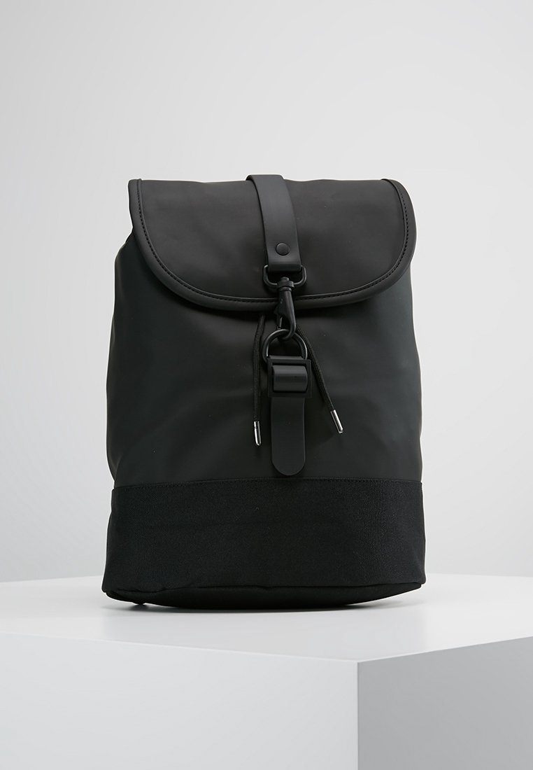 Sac à dos noir avec fermeture à rabat, sangle réglable et cordon sur un socle blanc, sur un fond clair uni.