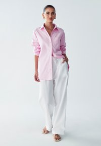 Camicia button-up rosa chiaro con maniche lunghe e taschino sul petto, abbinata a pantaloni bianchi larghi e sandali chiari. Design minimalista.
