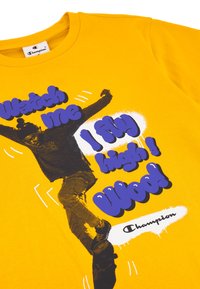 Žluté bavlněné tričko s černou grafikou osoby ve skoku a tučným fialovým textem "Dívej se, jak létám vysoko! Hurá!" s logem Champion.