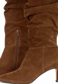 Bottes en suède marron jusqu'au genou avec un bout pointu, dotées d'une fermeture éclair sur le côté et de détails froncés le long de la tige ; accent en métal sur le talon.