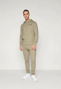 Khaki kapuce un joggers komplekts, kas izgatavots no mīksta materiāla, ar aukliņu kapuci, ribotām piedurknēm un melniem logotipa akcentiem. Valkāts ar baltām sporta apaviem.