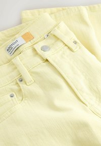 Jean bootcut jaune clair avec taille mi-haute, bouton métallique, passants pour ceinture et poches avant, étiqueté "Made in Pakistan".