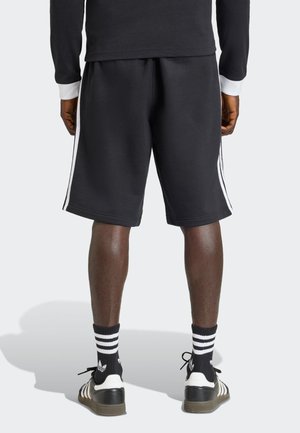 Čierne športové šortky so bielymi pruhmi na bokoch, vyrobené z mäkkého, štruktúrovaného materiálu. Kombinované s čiernymi a bielo pruhovanými ponožkami a teniskami.