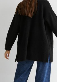 Černý pletený oversized svetr s dlouhými rukávy, bočními rozparky a texturovanou tkaninou, v kombinaci s modrými džíny.