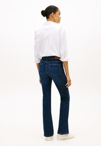 Mulher com cabelo apanhado em coque, vestindo camisa branca de botões, jeans azul escuro e sapatilhas brancas, em pé e olhando de lado contra um fundo branco.