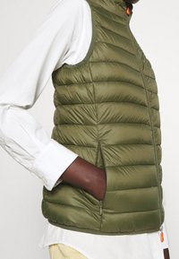 Olivgrön pufferväst med vertikalt quiltat mönster, hög krage och sidofickor med dragkedja. Tyget verkar slätt och lätt.