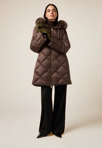 Brauner gesteppter Steppmantel mit Kunstpelzkapuze, vorne mit Reißverschluss und Taschen, kombiniert mit schwarzen, weit geschnittenen Hosen und grünen Handschuhen.