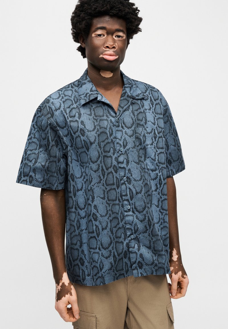 Camisa de manga corta con botones en estampado de serpiente azul y negro, que presenta un cuello, corte holgado y textura de tela suave.