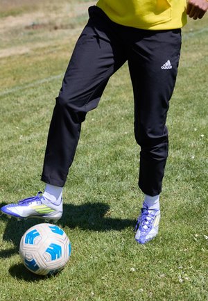 Osoba w czarnych spodniach Adidas i fioletowych korkach kontroluje białą i niebieską piłkę nożną na zielonym trawiastym boisku.