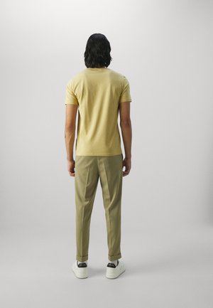 Mann mit schwarzen, welligen Haaren, der ein senffarbenes kurzärmeliges Hemd, beige Hosen und weiße Turnschuhe trägt und mit dem Rücken zur Kamera steht.